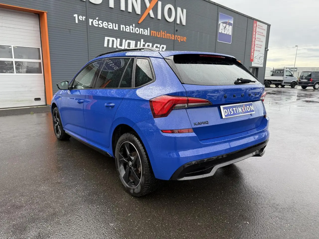 Vue 3/4 arrière gauche d'un Skoda Kamiq Bleu Energy avec jantes alliage noires et vitres surteintées sous la pluie.
