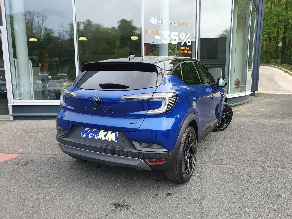 Vue 3/4 arrière droite d’un Renault Captur bleu Iron avec jantes noires et vitres surteintées, stationné devant un bâtiment.