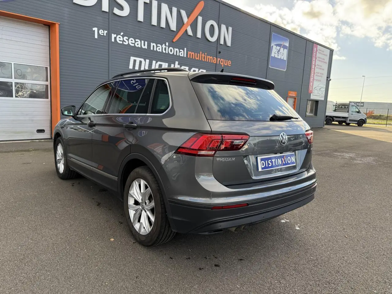 Vue 3/4 arrière droite d'un Volkswagen Tiguan gris Indium avec vitres arrière surteintées, garé devant un bâtiment commercial.
