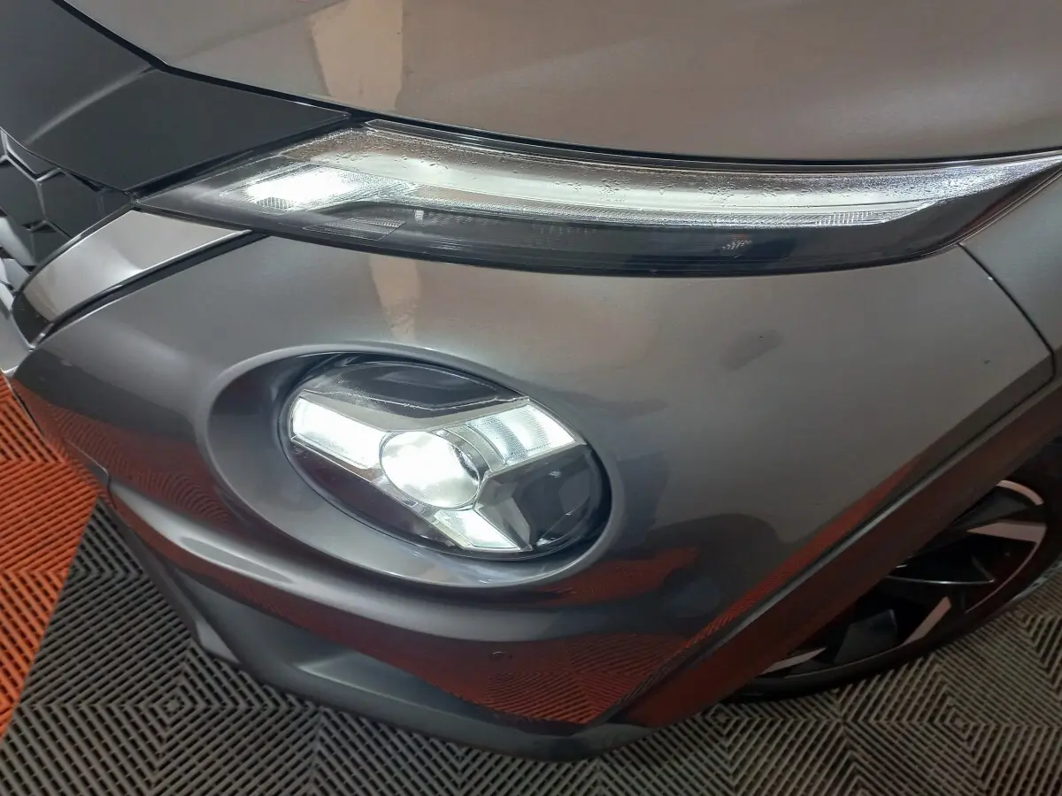 Gros plan sur le phare avant droit allumé d'un Nissan Juke Hybrid 143 Business+ gris foncé, avec partie de la calandre visible.