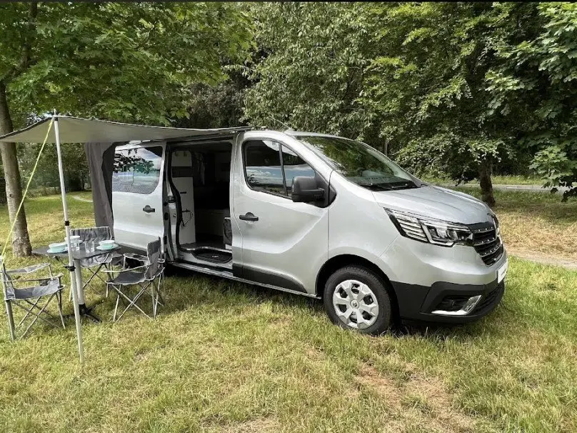 Renault Trafic Fourgon gris vu en 3/4 avant droit, porte latérale ouverte avec auvent et chaises de camping installées.