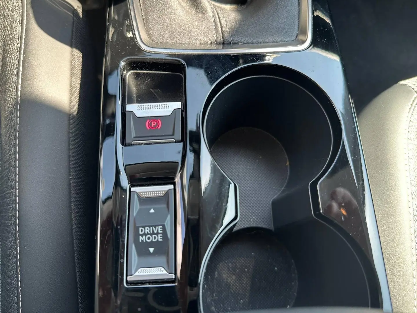Gros plan sur la console centrale noire brillante de la Peugeot 208 1.2 100ch, avec commandes frein à main électrique et drive mode.