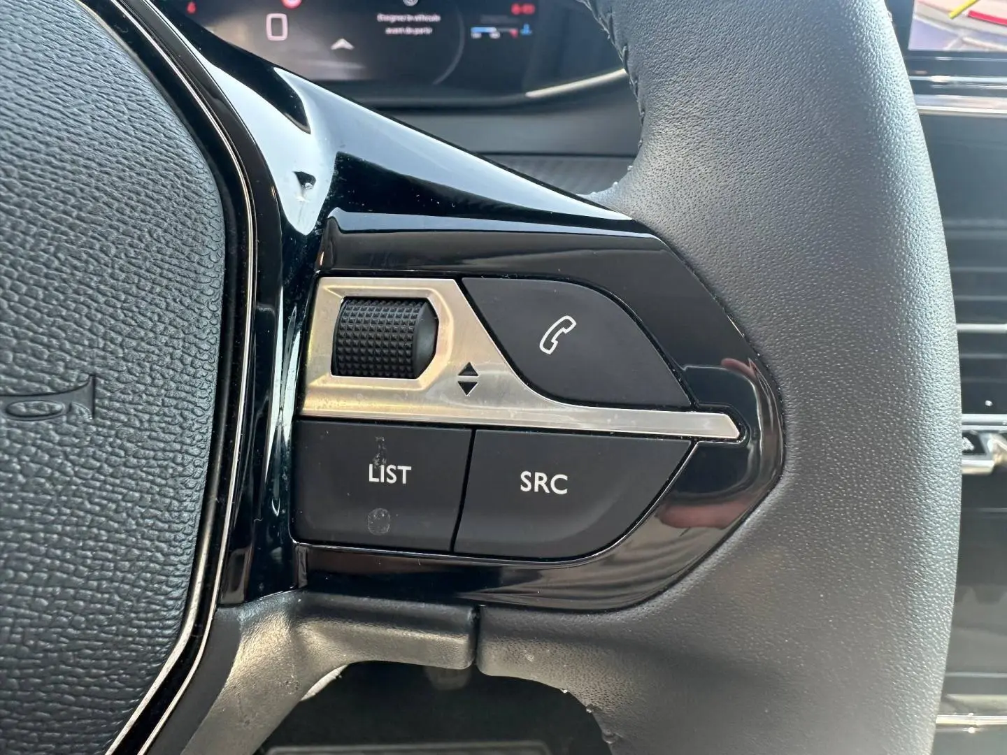 Gros plan sur les commandes gauche du volant noir de la Peugeot 208 2025, boutons SRC, LIST et molette de réglage.