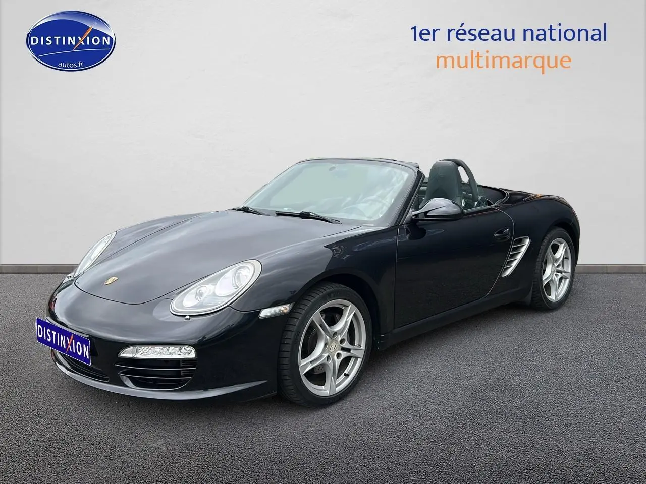 Vue 3/4 avant droite d'une Porsche Boxster 987 noire cabriolet avec jantes alu 17 pouces sur sol gris.