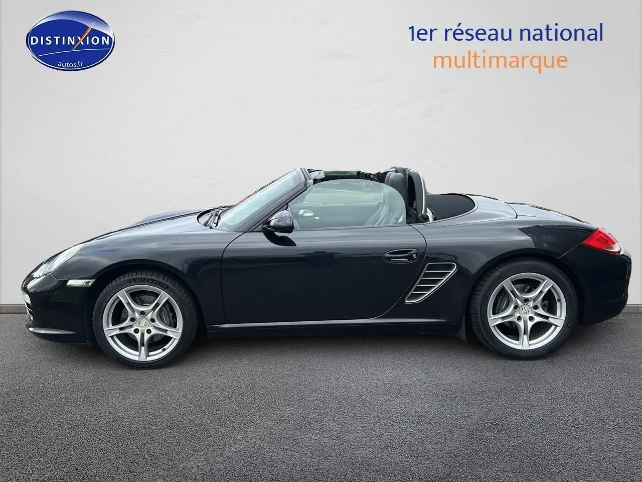 Profil côté gauche d'une Porsche Boxster 987 noire cabriolet avec jantes alu 17 pouces sur fond clair.