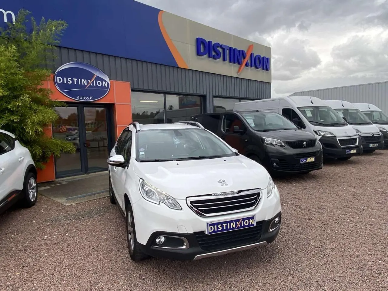 Peugeot 2008 blanc en 3/4 avant droit devant un concessionnaire Distinxion sous un ciel nuageux.
