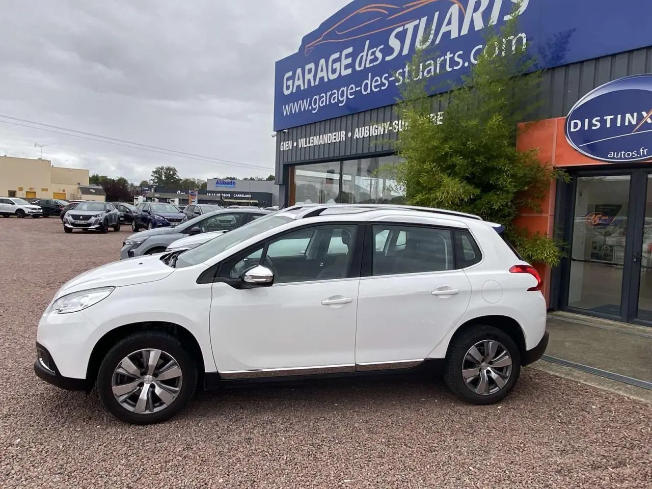 Profil côté gauche d'un Peugeot 2008 blanc 2015 garé devant un garage, avec jantes alliage et rétroviseurs chromés.