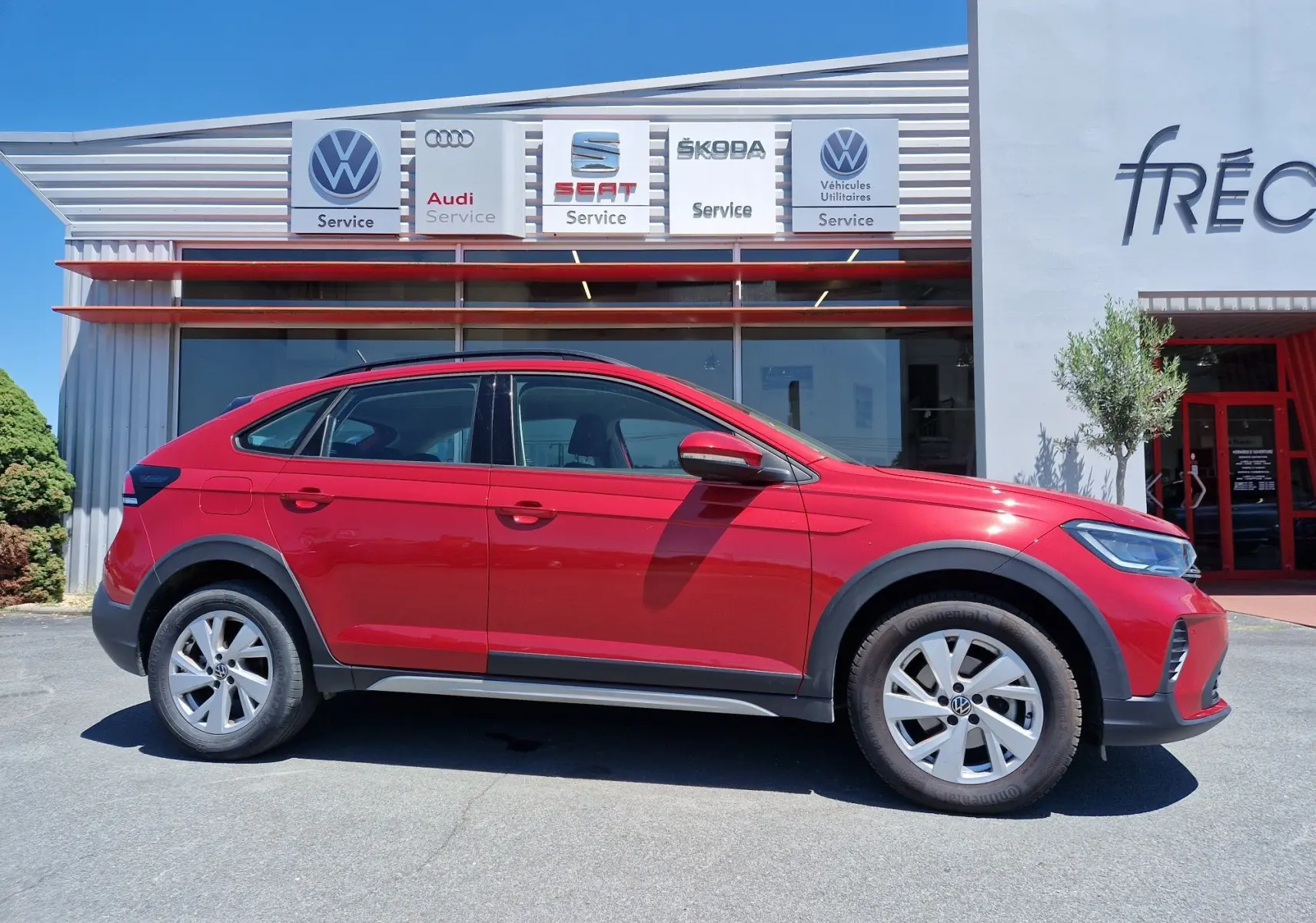 Vue de profil côté gauche d'un Volkswagen Taigo rouge roi garé devant un service auto multimarques.