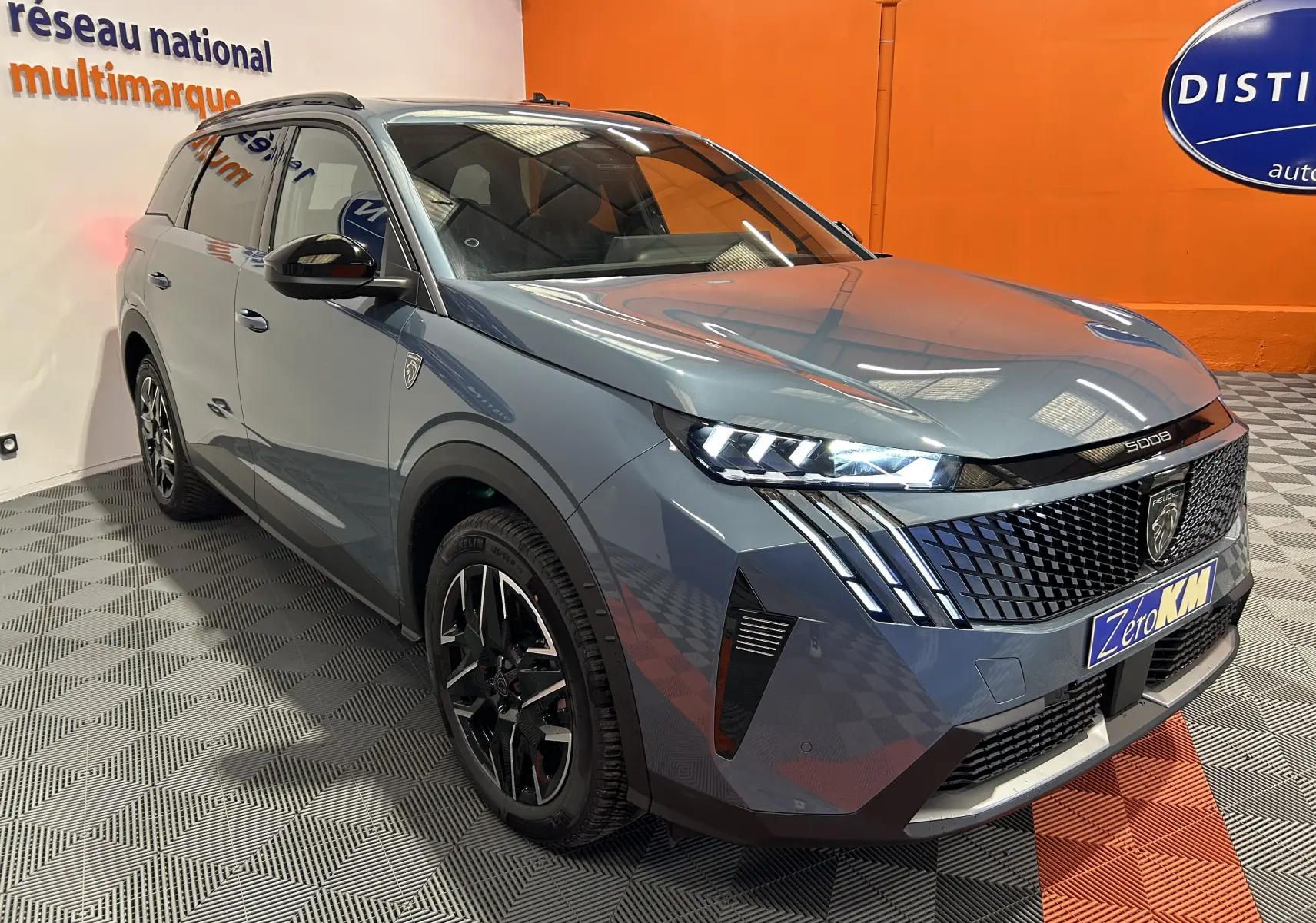 Vue 3/4 avant droit d’un Peugeot 5008 bleu Ingaro avec phares LED et toit ouvrant panoramique en intérieur showroom.