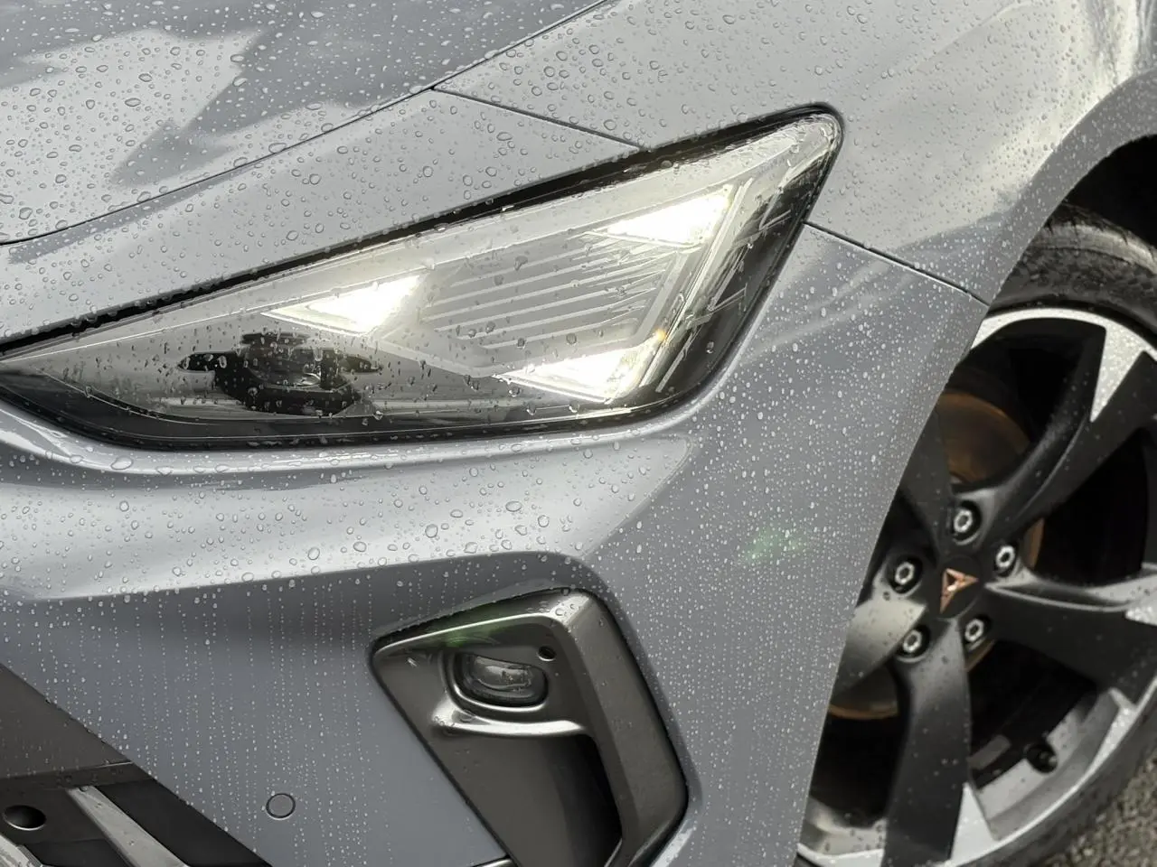 Gros plan sur le phare avant droit allumé et la jante noire de la CUPRA LEON ST gris Graphene sous la pluie.