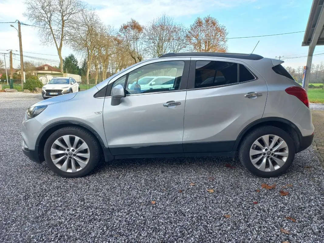 Vue de profil côté gauche d'un Opel Mokka X gris 2017 garé sur un sol gravillonné avec des jantes alliage.