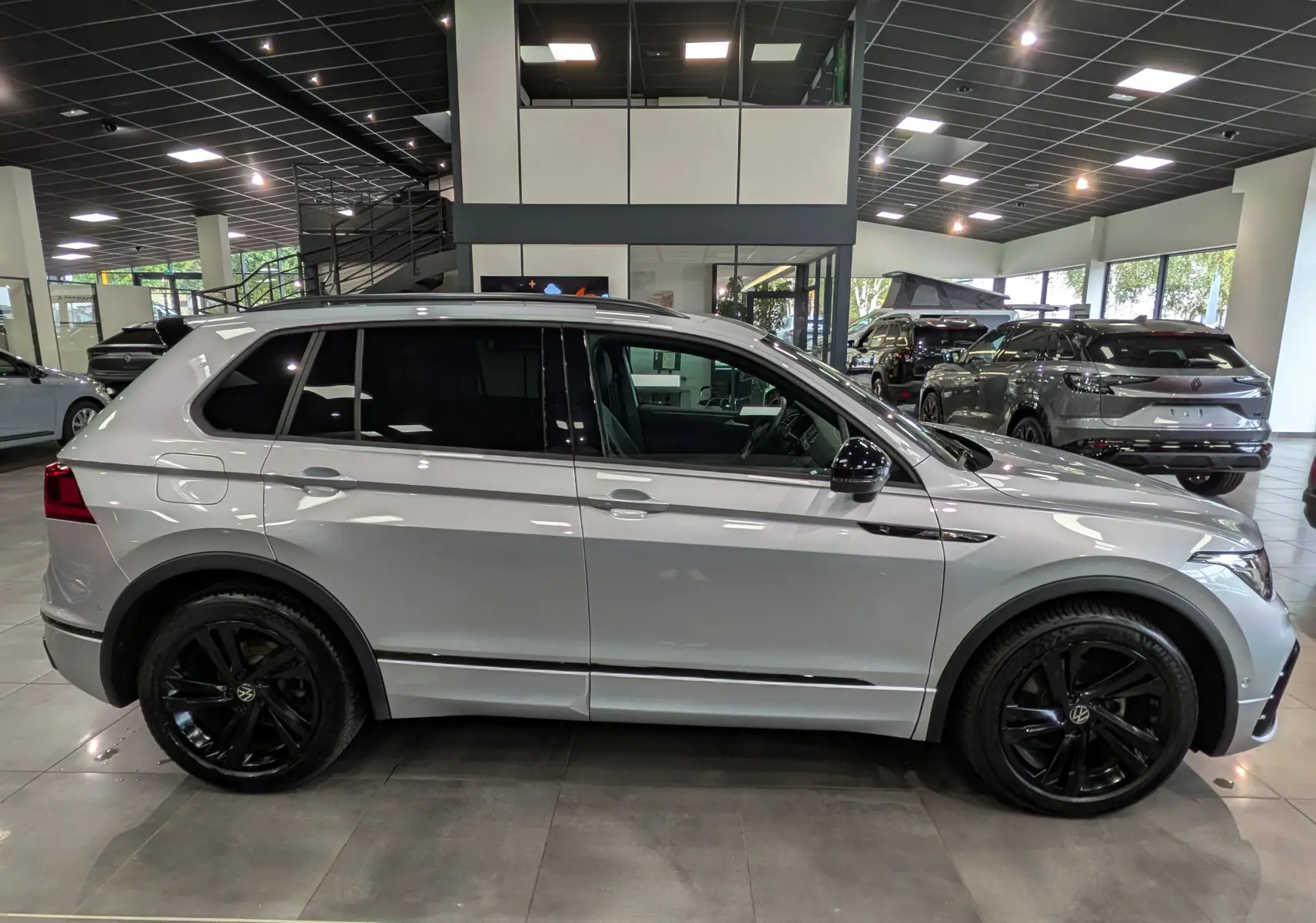 Vue de profil côté gauche d’un Volkswagen Tiguan 2024 reflet d’argent avec jantes noires et vitres teintées en showroom.