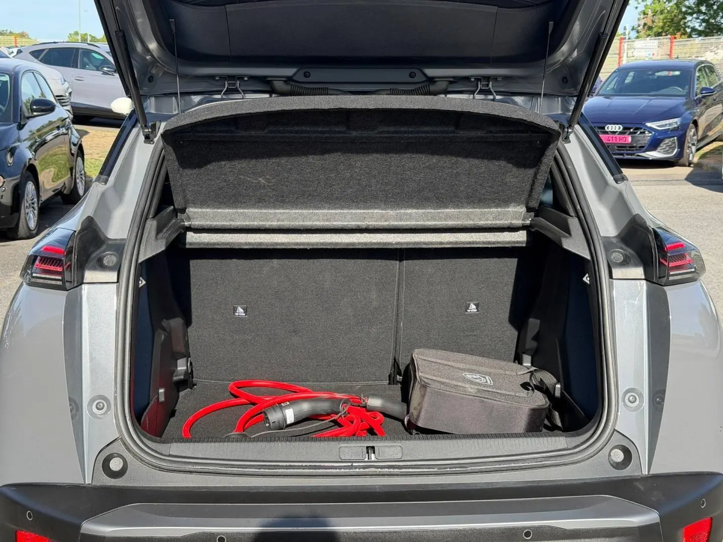 Coffre ouvert d’un Peugeot e-2008 gris Artense, vue arrière, avec câble de recharge électrique rouge visible.