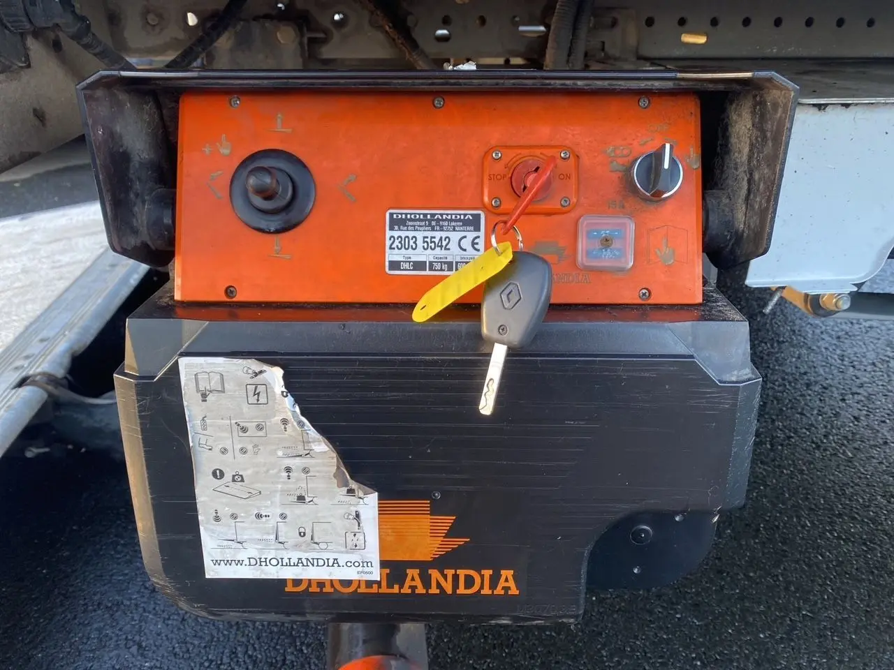 Gros plan sur le boîtier orange du hayon élévateur Dhollandia avec clé Renault accrochée dessous du châssis blanc.
