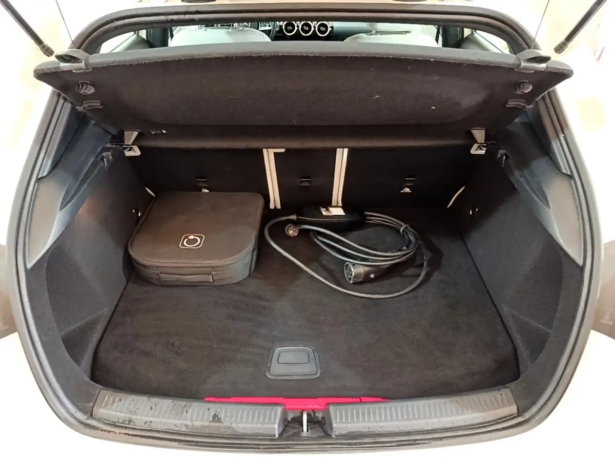 Vue intérieure du coffre ouvert d'une Mercedes Classe A Berline blanche, avec câble de recharge et sac de rangement.