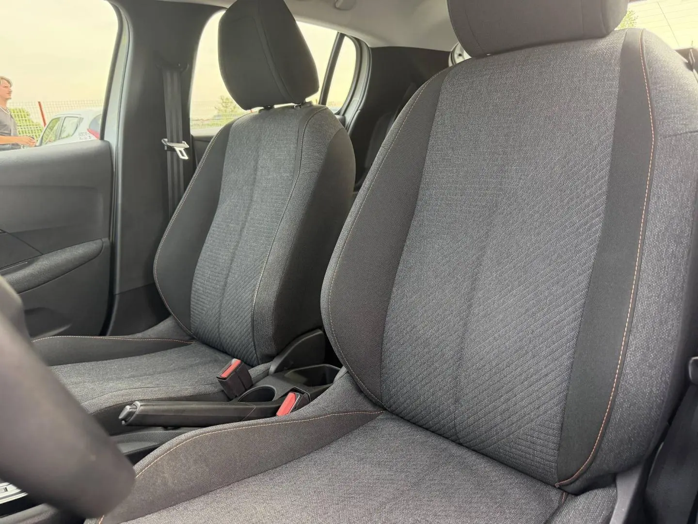 Vue rapprochée des sièges avant en tissu gris avec surpiqûres orange de la Peugeot 208 hybride blanche.