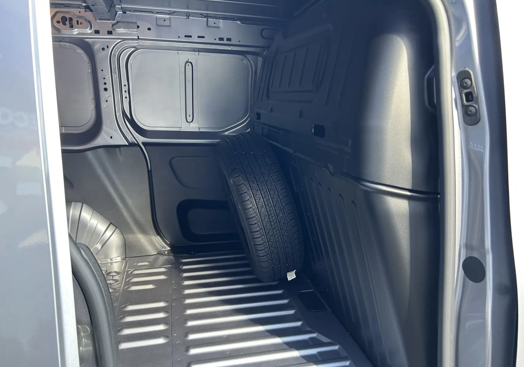 Intérieur de la zone de chargement côté droit du DANGEL Partner gris Artense avec une roue de secours appuyée contre la paroi.