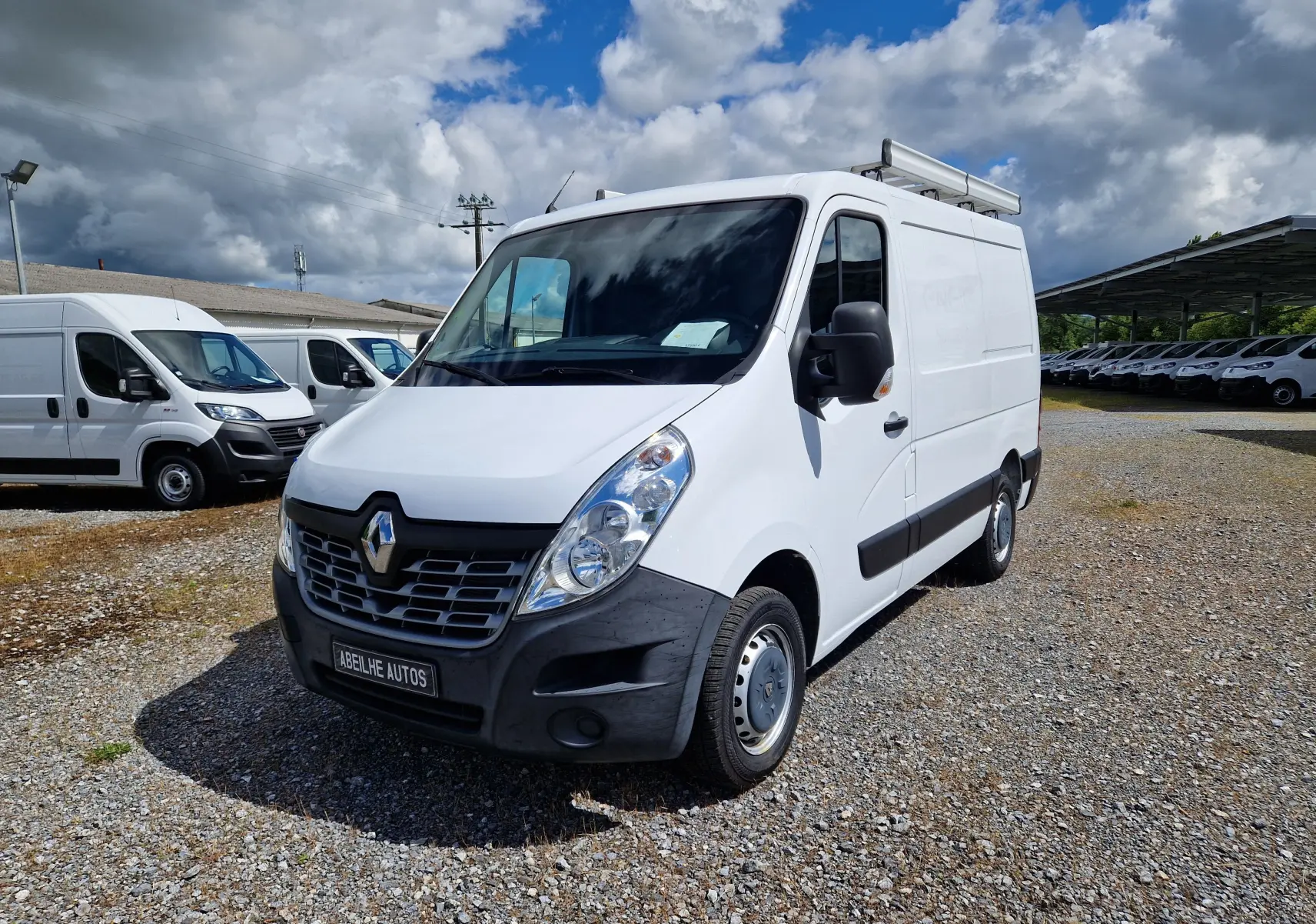 Renault Master blanc en vue 3/4 avant droit avec galerie de toit, stationné sur un sol gravillonné sous ciel nuageux.
