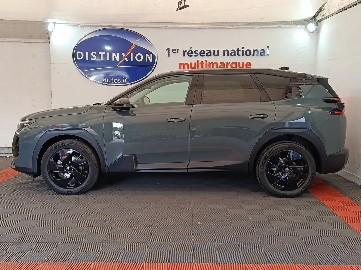 Vue de profil côté gauche d'une Citroën C5 hybride 2026 vert foncé avec jantes noires brillantes en intérieur showroom.