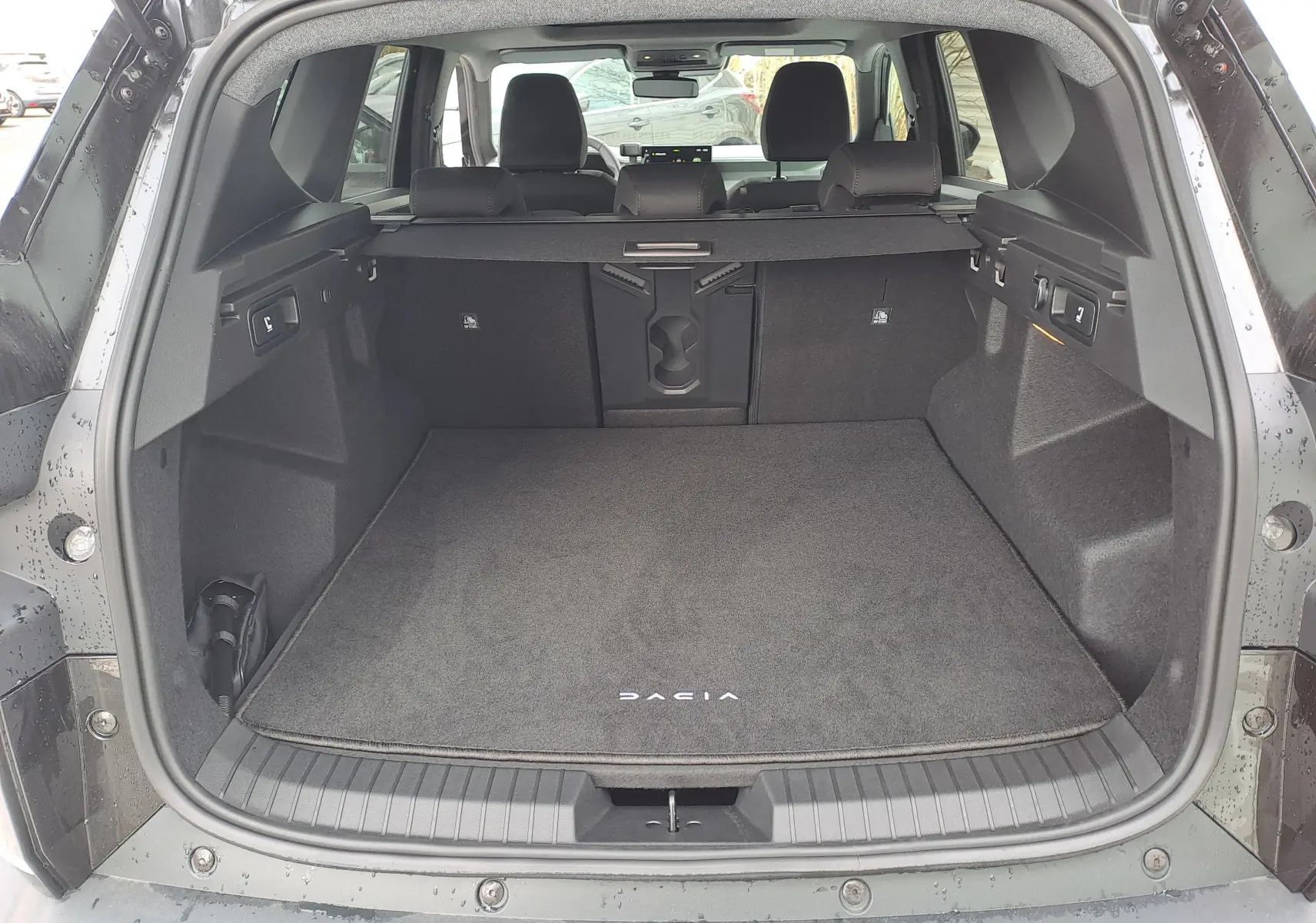 Coffre ouvert vu de l'arrière d'un Dacia Bigster noir nacré, avec tapis de coffre marqué Dacia et banquette arrière visible.