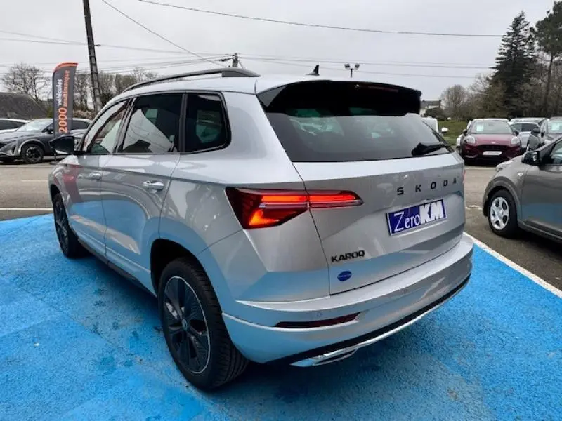 Vue 3/4 arrière droite d’un Skoda Karoq gris diamant métallisé avec feux arrière LED allumés sur parking.