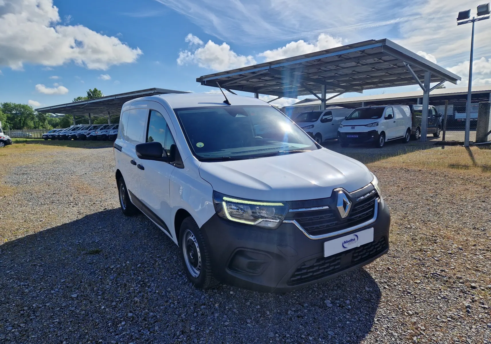 Renault Kangoo blanc en 3/4 avant droit sur parking gravillonné, avec calandre noire et phares allumés.
