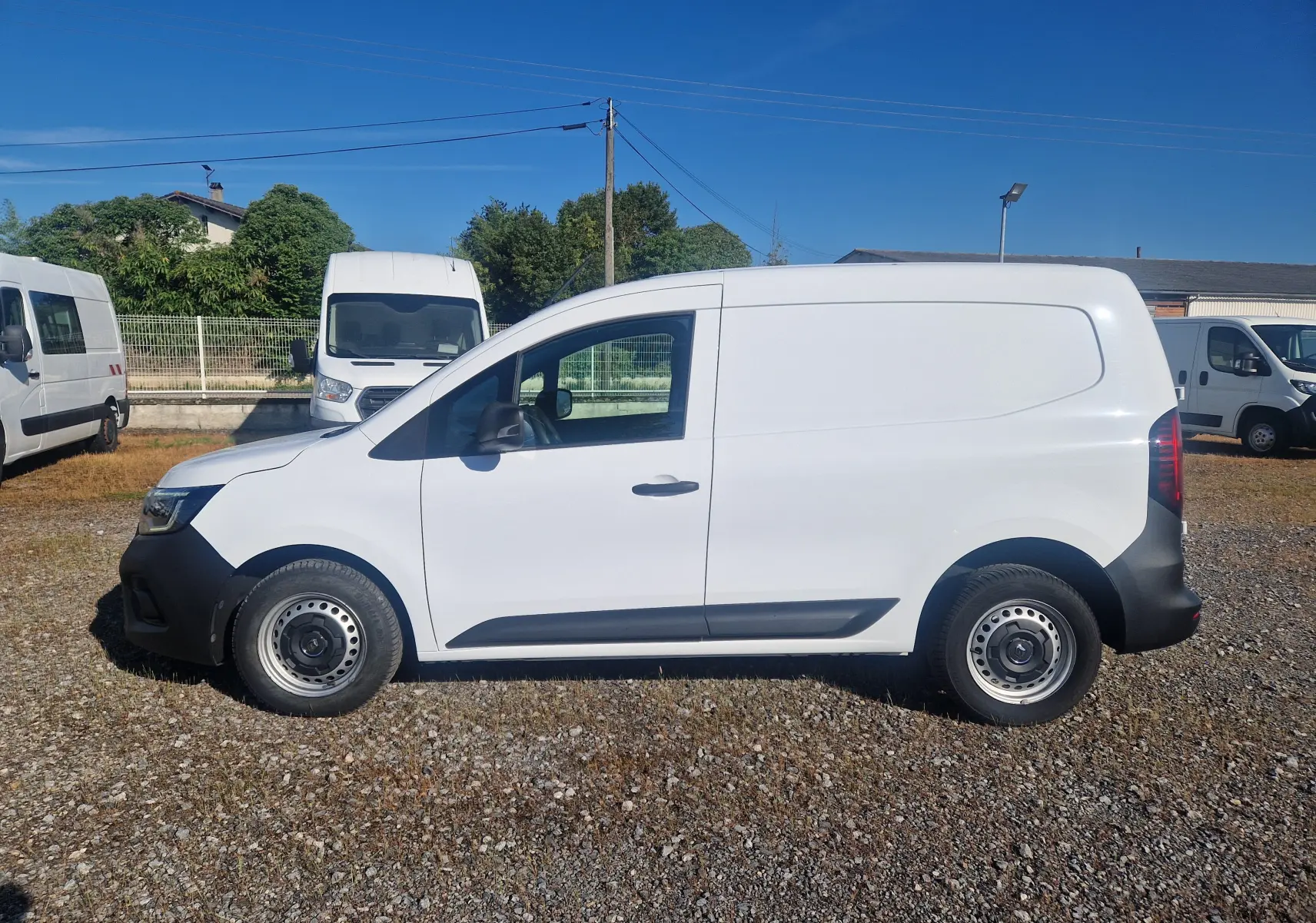 Vue de profil côté gauche d'un Renault Kangoo blanc 2023 utilitaire avec protections noires sur un terrain gravillonné.