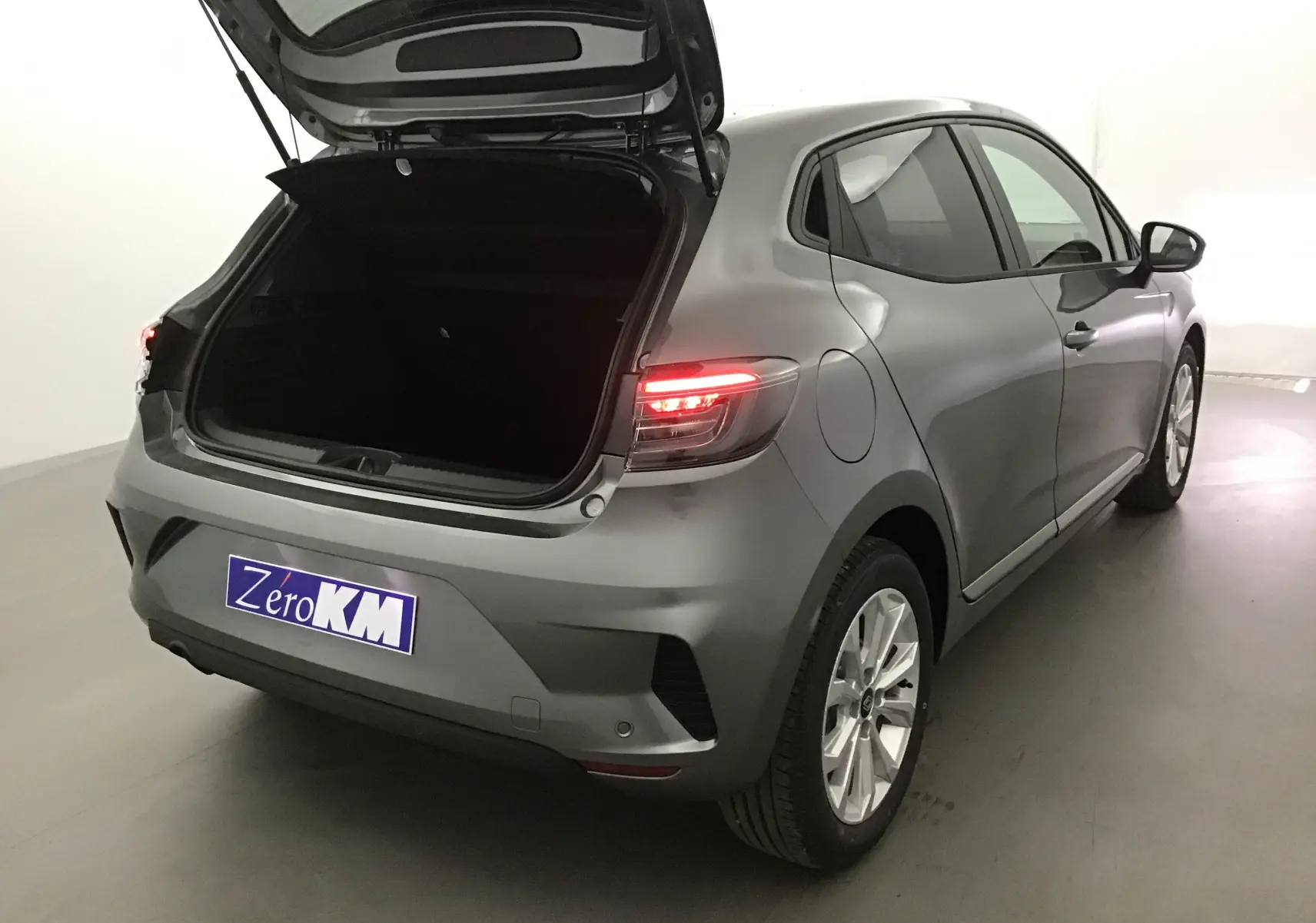 Vue 3/4 arrière droite d'une Renault Clio V gris schiste avec coffre ouvert et feux arrière allumés.