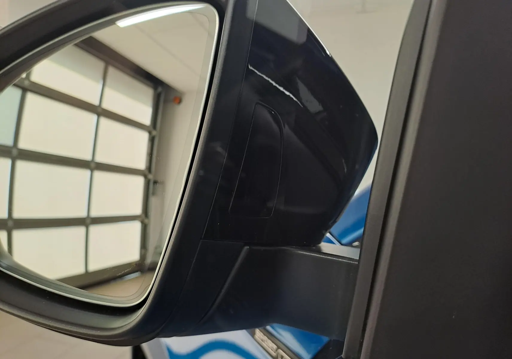 Gros plan sur le rétroviseur côté conducteur noir brillant du Skoda Karoq Bleu RS 2025 avec reflet de la porte de garage.