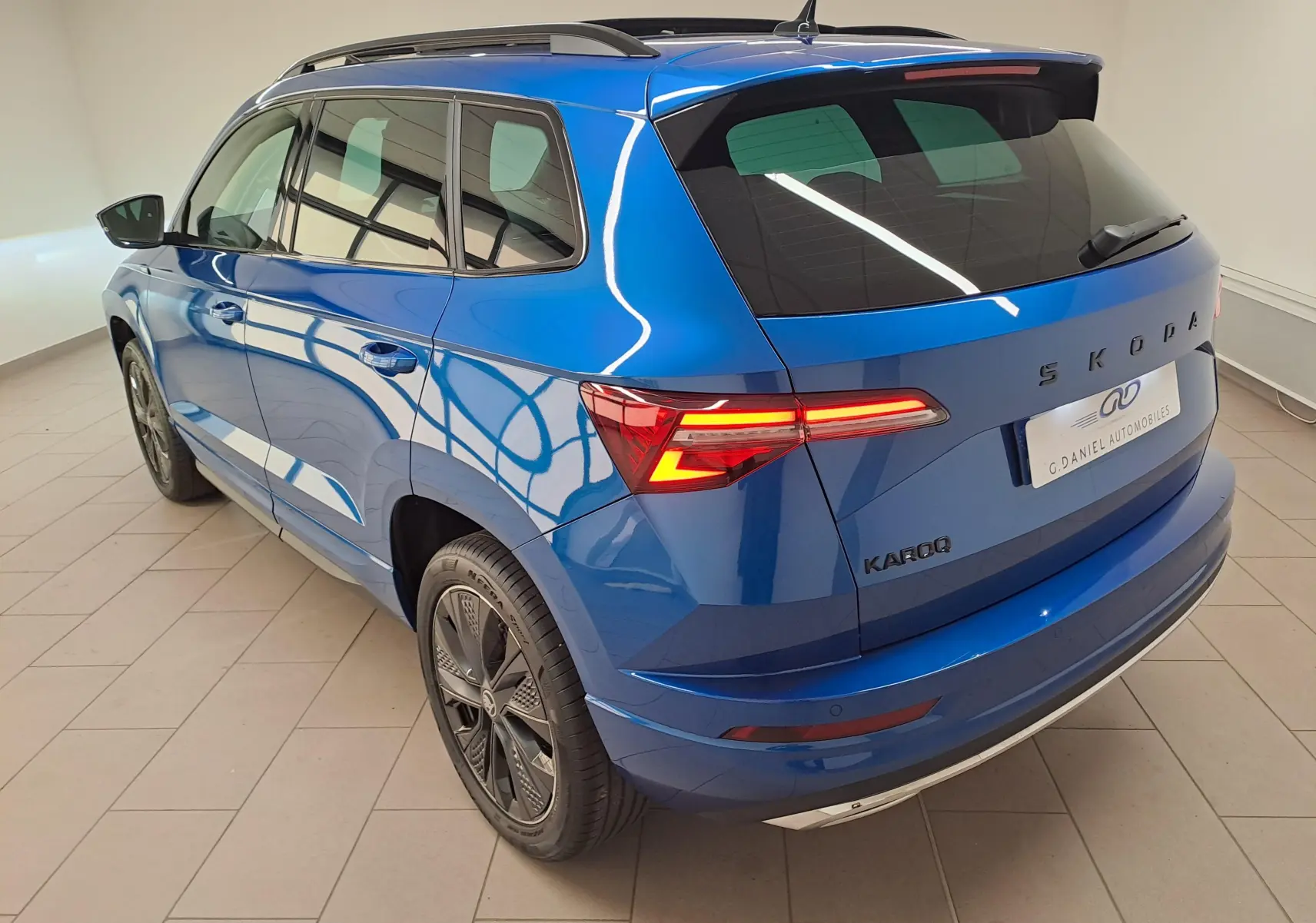 Vue 3/4 arrière gauche d'un Skoda Karoq bleu RS avec feux allumés et toit panoramique ouvrant visible.