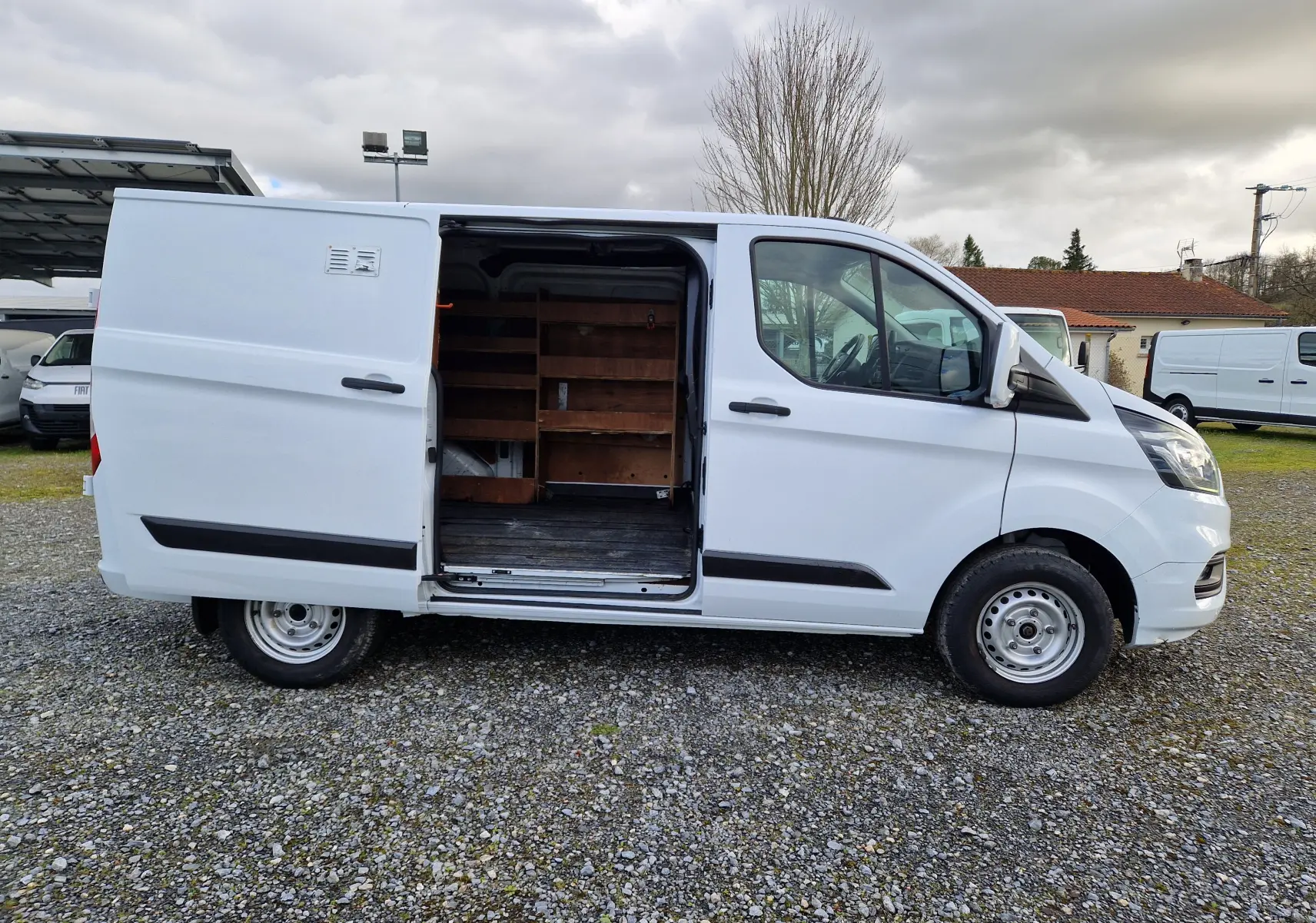 Ford Transit Custom Fourgon blanc, vue de profil côté gauche sur un sol gravillonné, sans fenêtres latérales arrière.