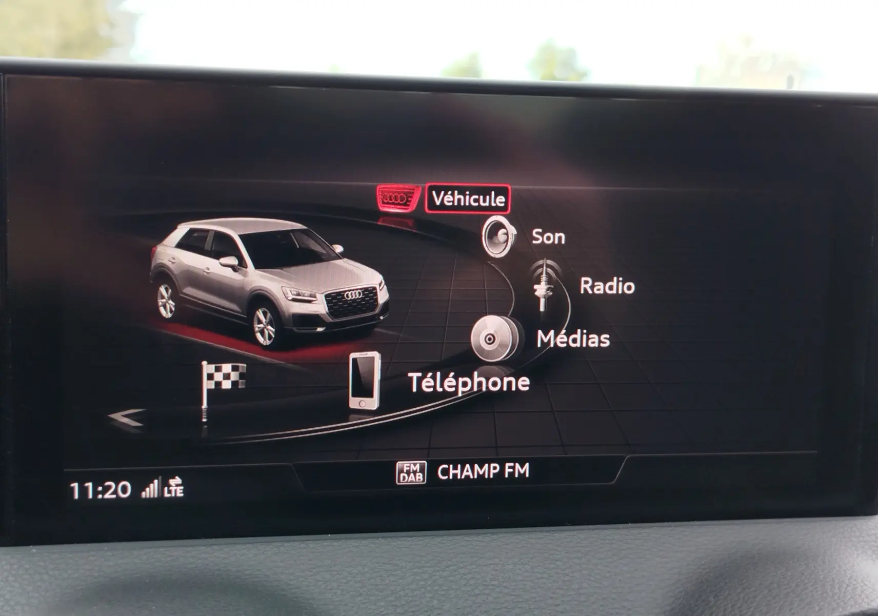Écran multimédia affichant une Audi Q2 grise en vue 3/4 avant avec menu de navigation et options audio visibles.