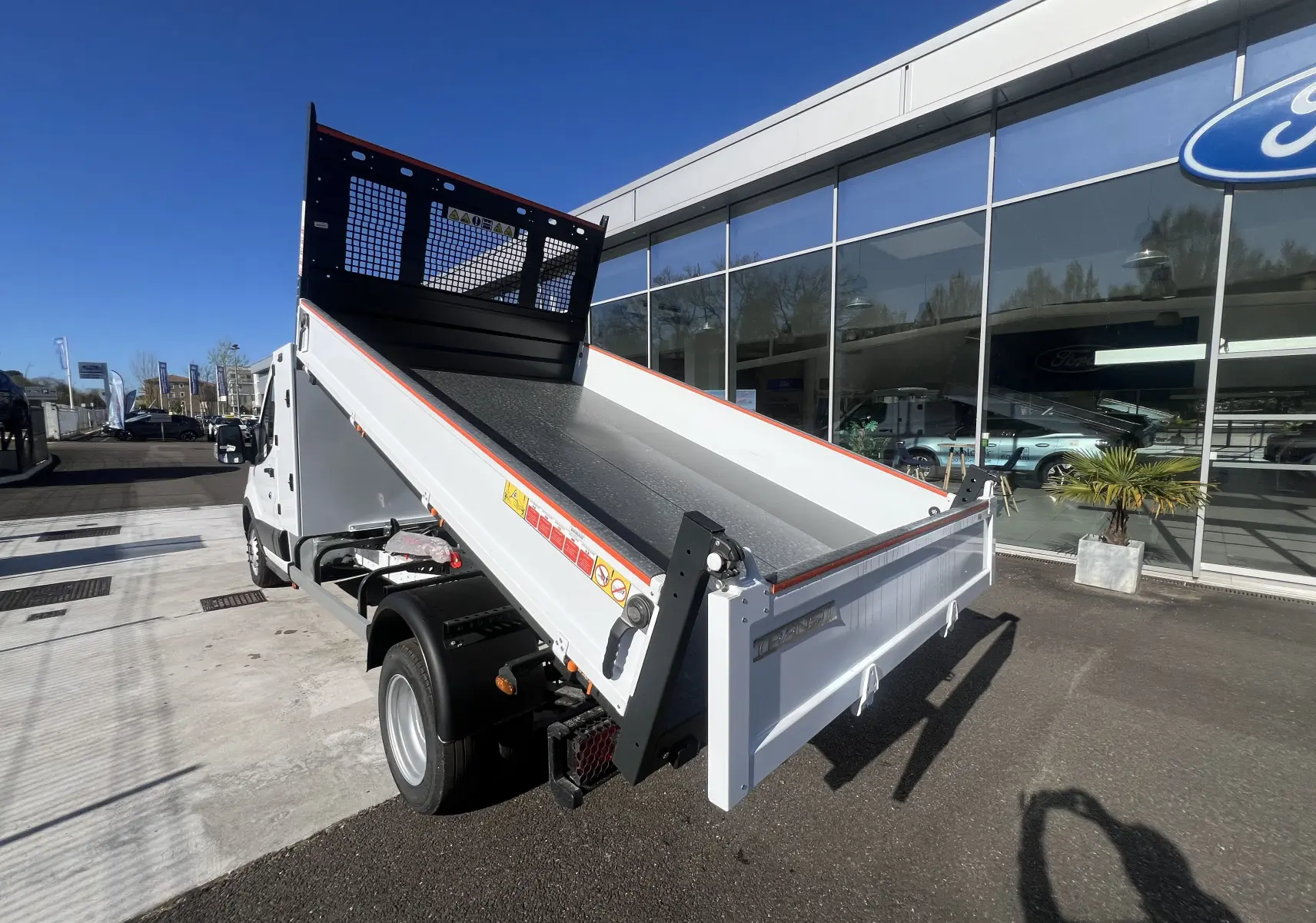 Vue 3/4 arrière droite du Ford Transit Chassis Cabine blanc avec benne relevée et coffre, stationné devant un bâtiment Ford.