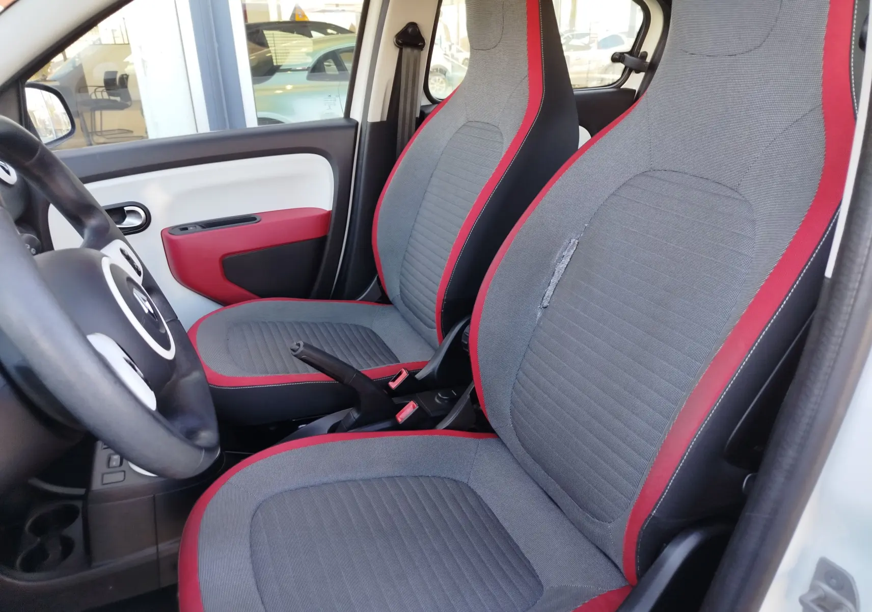 Vue intérieure côté gauche de la Renault Twingo 1.0 SCe 70ch Zen 2016 avec sièges gris et rouge et portière blanche.