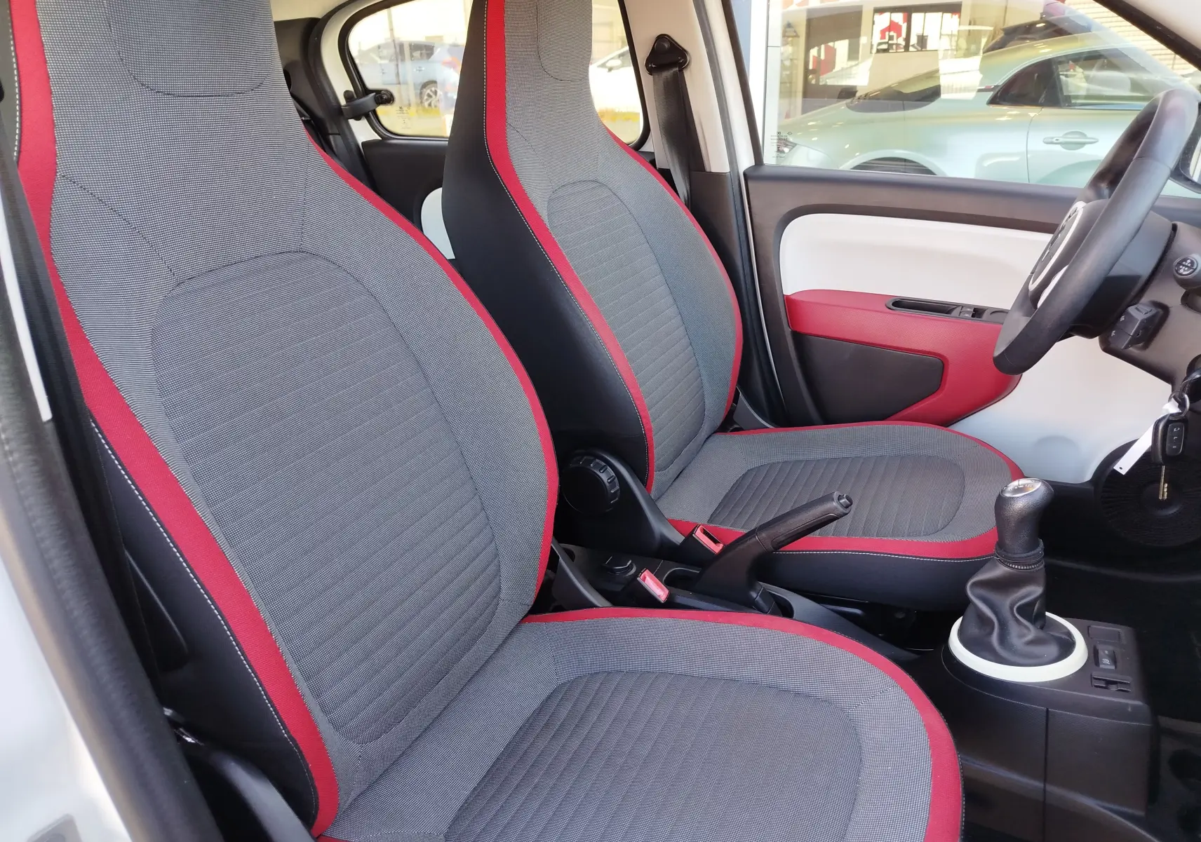 Vue intérieure côté conducteur de la Renault Twingo 2016, sièges gris avec contours rouges et volant à droite.