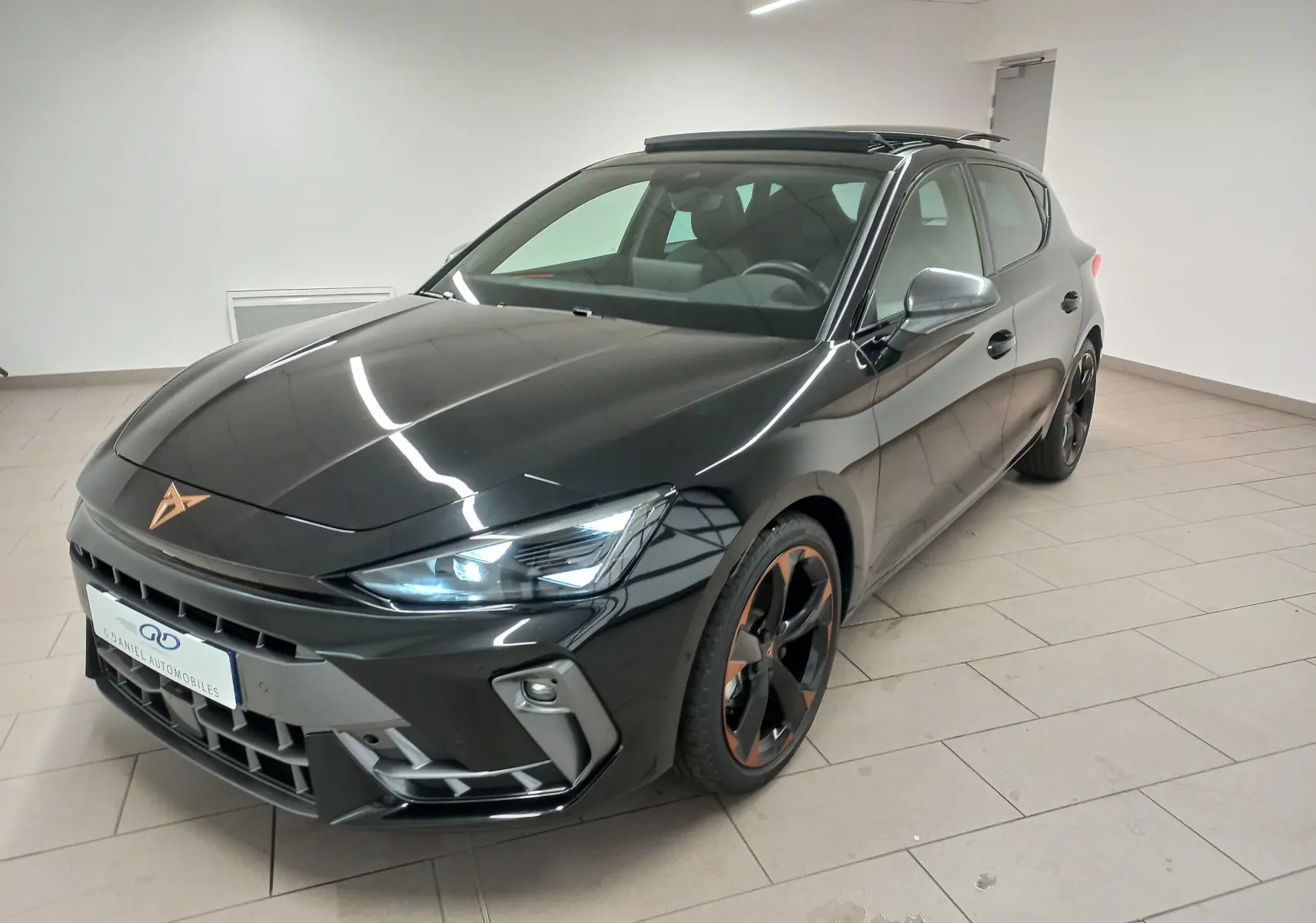 Vue 3/4 avant droite d'une CUPRA Leon noire Minuit avec jantes cuivrées et toit ouvrant panoramique en intérieur showroom.