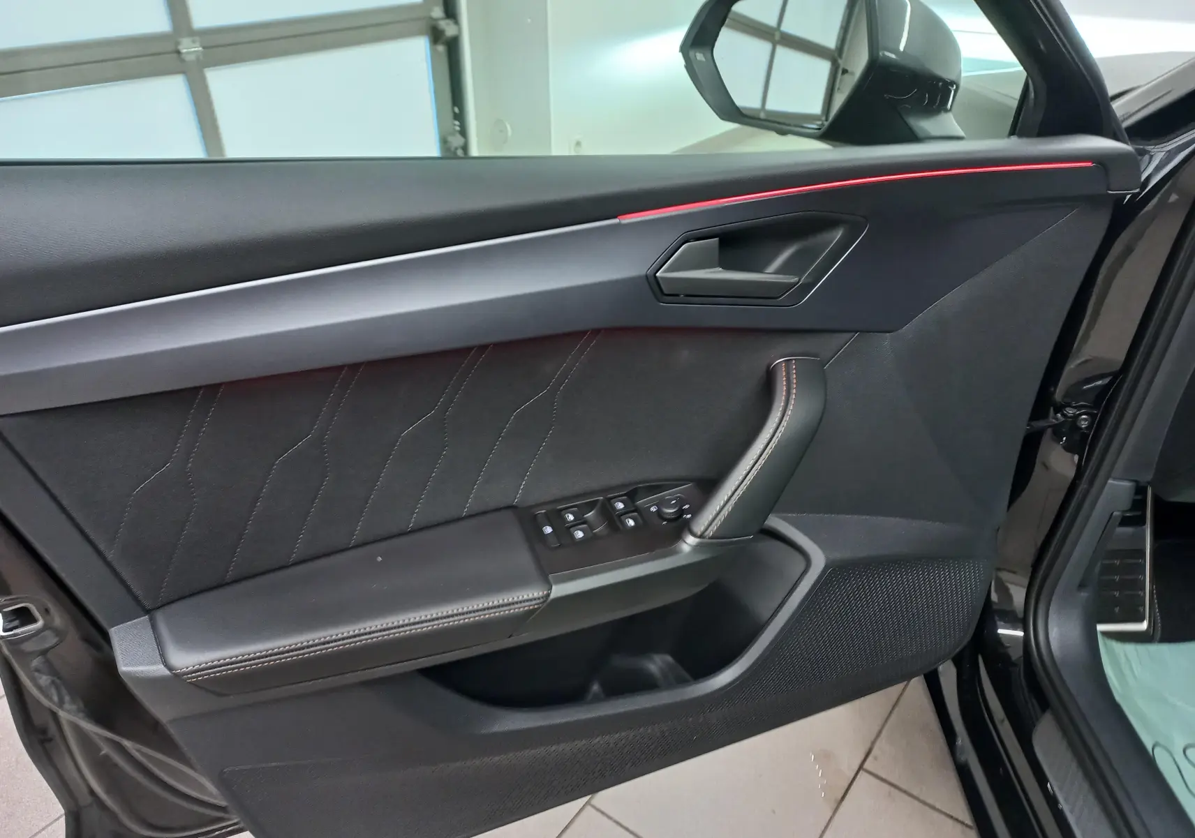 Vue intérieure côté gauche de la porte d'une CUPRA Leon 2025 noir minuit avec surpiqûres cuivrées et bande lumineuse rouge.
