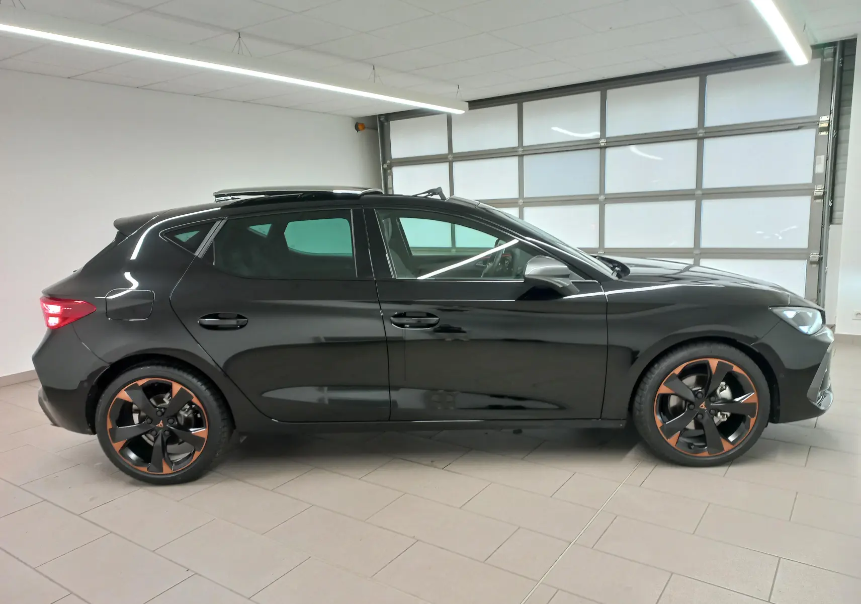 Profil droit d'une CUPRA Leon 2025 noir minuit avec toit ouvrant panoramique et jantes cuivrées 18 pouces en intérieur showroom.