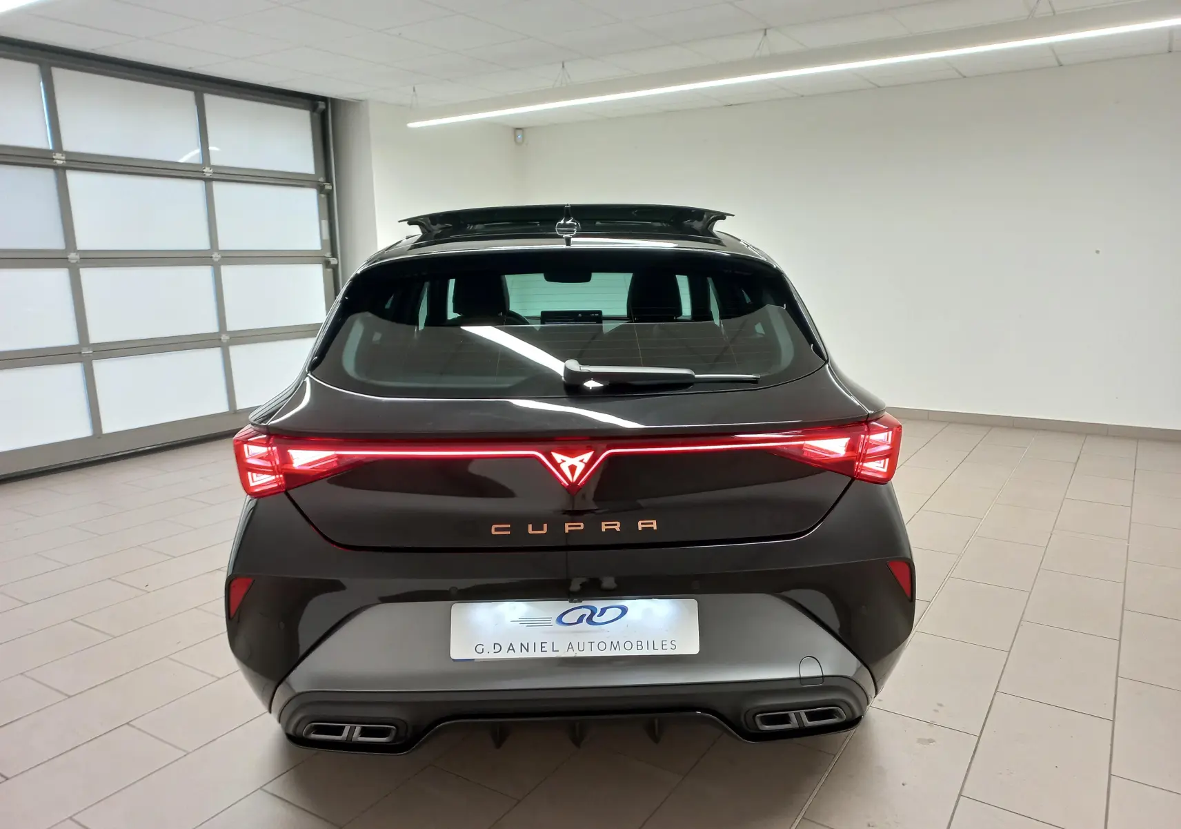 Vue arrière d'une CUPRA Leon 2025 Noir Minuit avec feux LED allumés et logo cuivré dans un garage blanc.