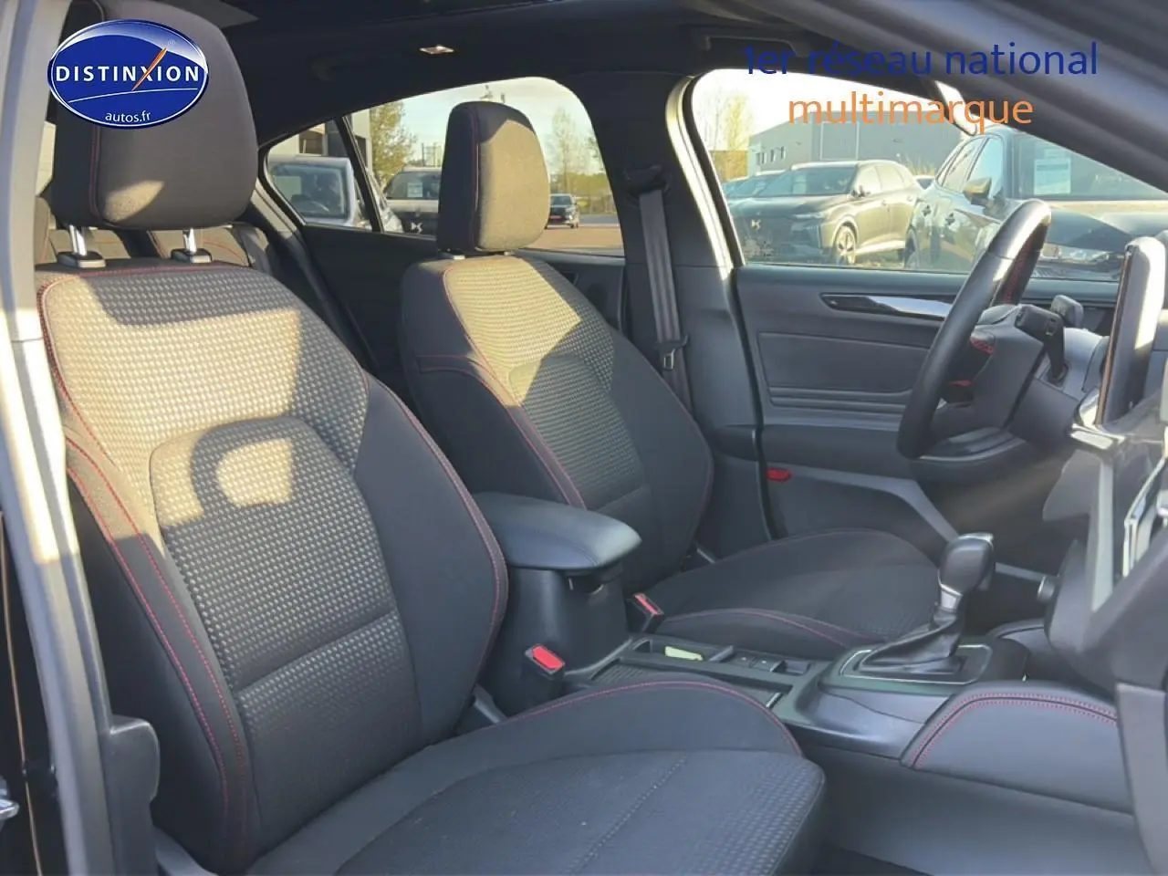 Vue intérieure avant droite de la Ford Focus noire 2024 avec sièges tissu noir et surpiqûres rouges, console centrale et volant cuir.
