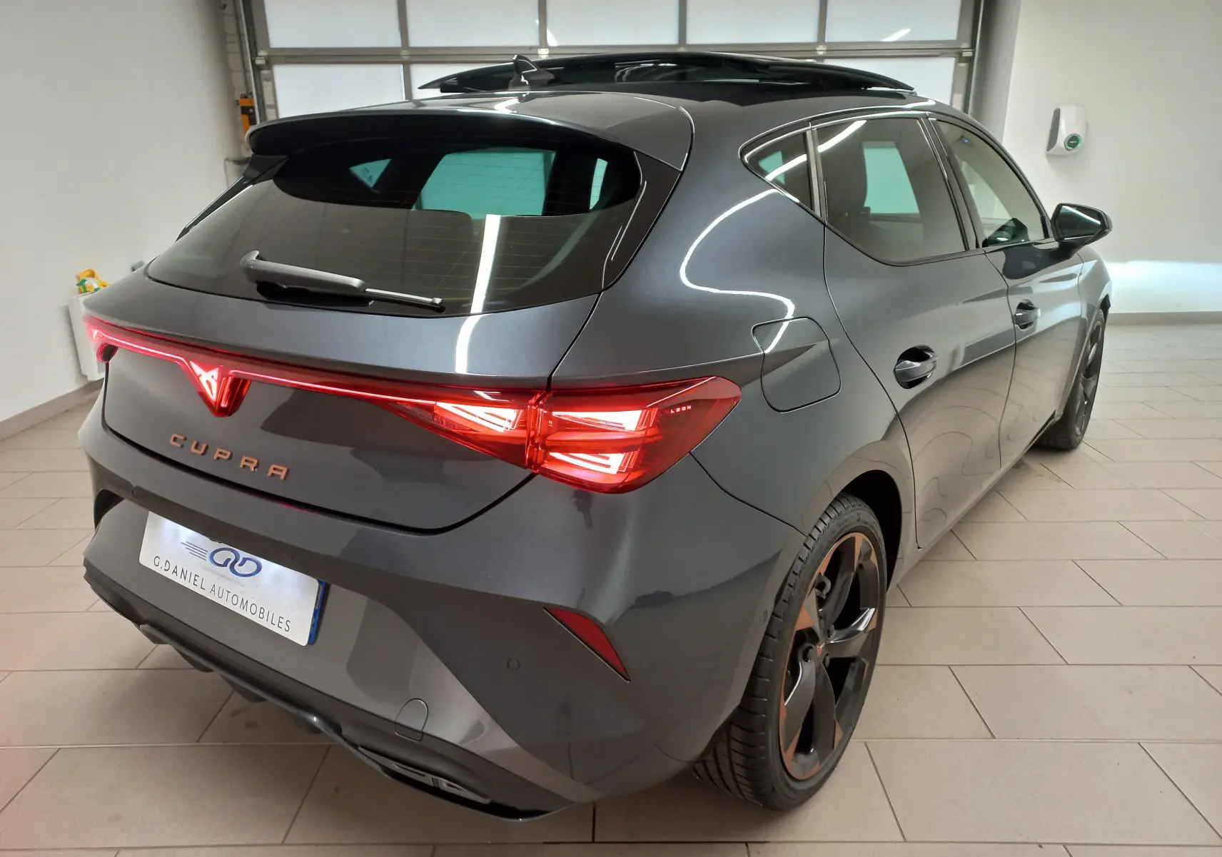 Vue 3/4 arrière droite d'une CUPRA Leon gris magnétique avec jantes cuivrées et toit ouvrant panoramique en intérieur.