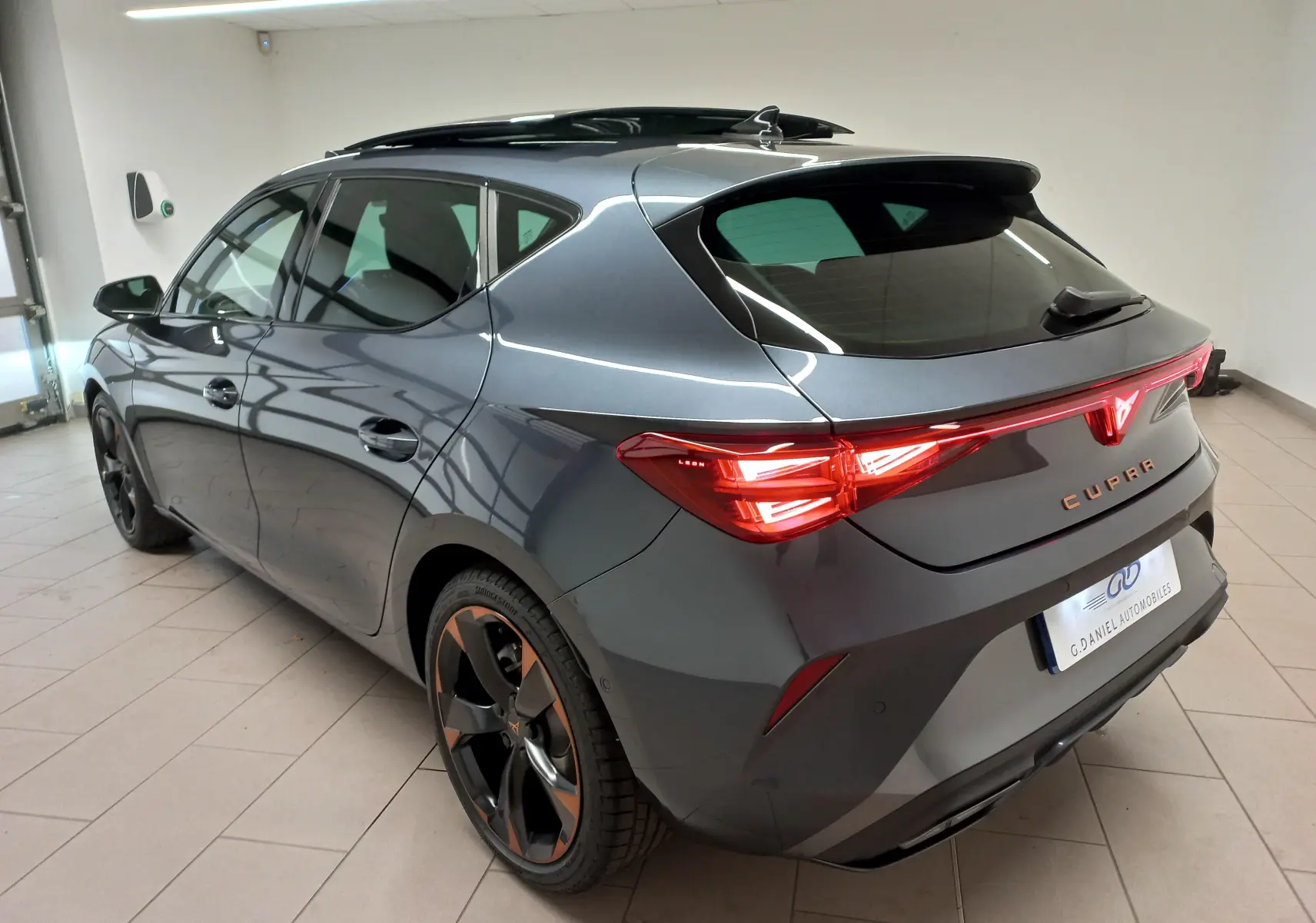 Vue 3/4 arrière droite d'une CUPRA Leon gris magnétique avec jantes cuivrées et toit ouvrant panoramique en intérieur.