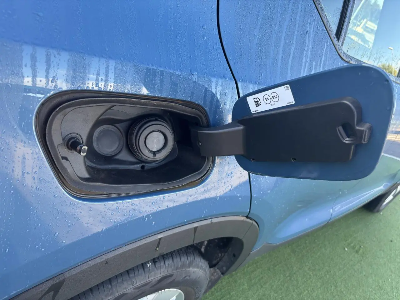 Gros plan sur la trappe à carburant ouverte côté gauche d'un Volvo XC40 Fjord Blue Metal avec gouttes d'eau.