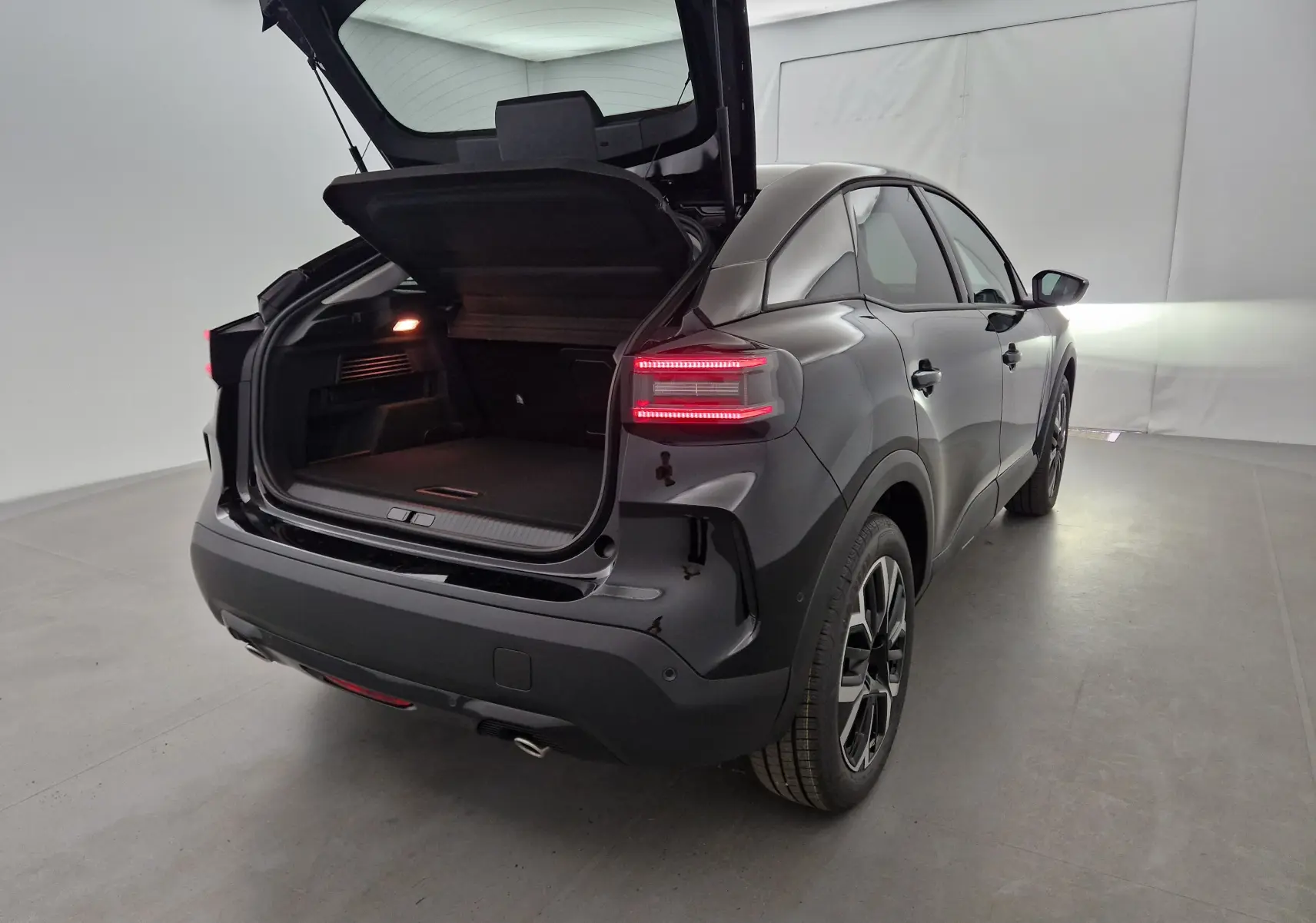 Vue 3/4 arrière droite d'une Citroën C4 noire 2025 avec coffre ouvert et feux arrière allumés dans un studio lumineux.