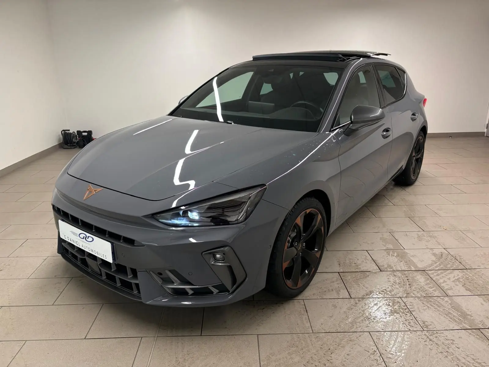 Vue 3/4 avant droit d'une CUPRA Leon gris Graphène avec toit ouvrant panoramique et jantes alliage cuivrées en intérieur showroom.
