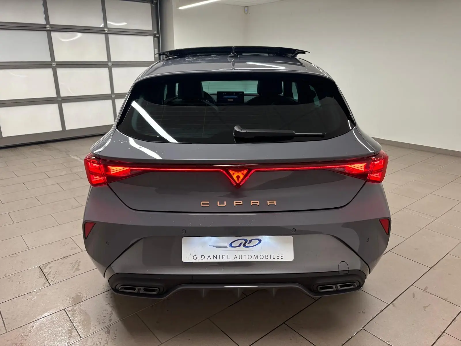 Vue arrière d'une CUPRA Leon gris Graphène avec feux LED allumés et toit ouvrant panoramique visible en intérieur showroom.