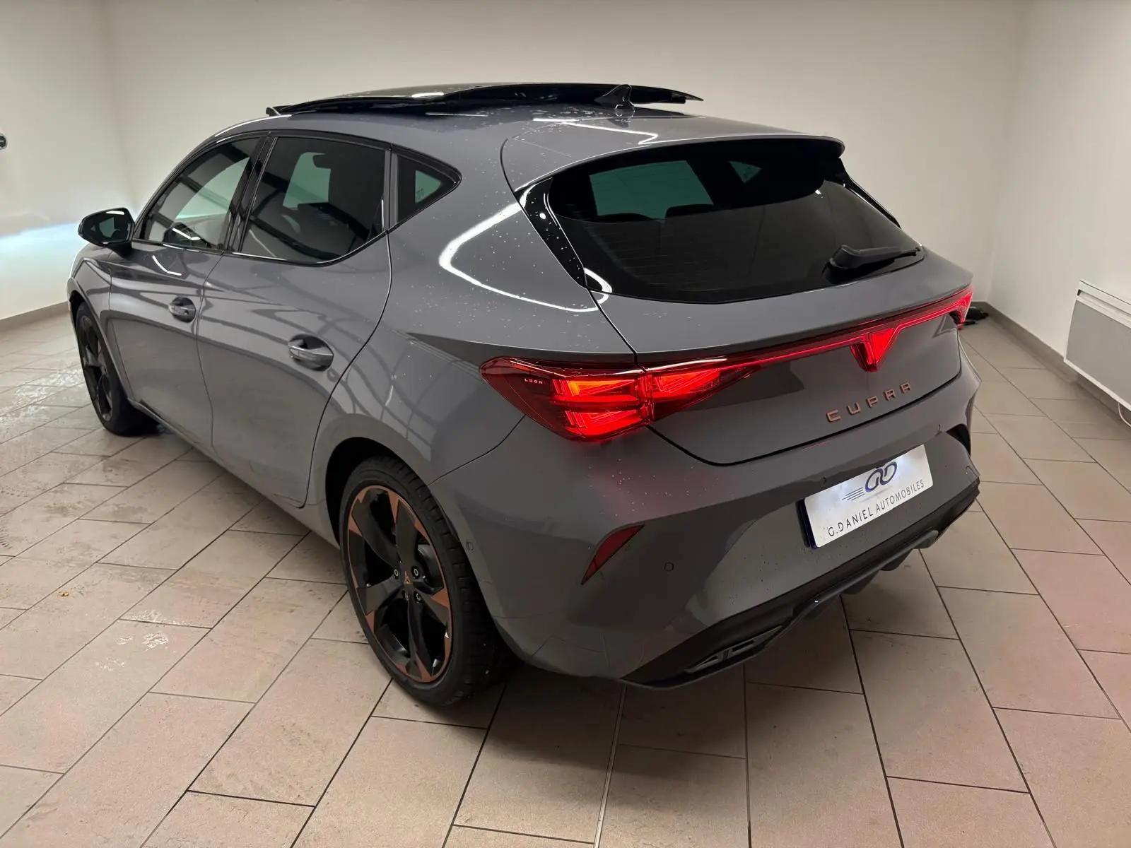 Vue 3/4 arrière droite d'une CUPRA Leon gris Graphène avec jantes cuivrées et toit ouvrant panoramique en intérieur.