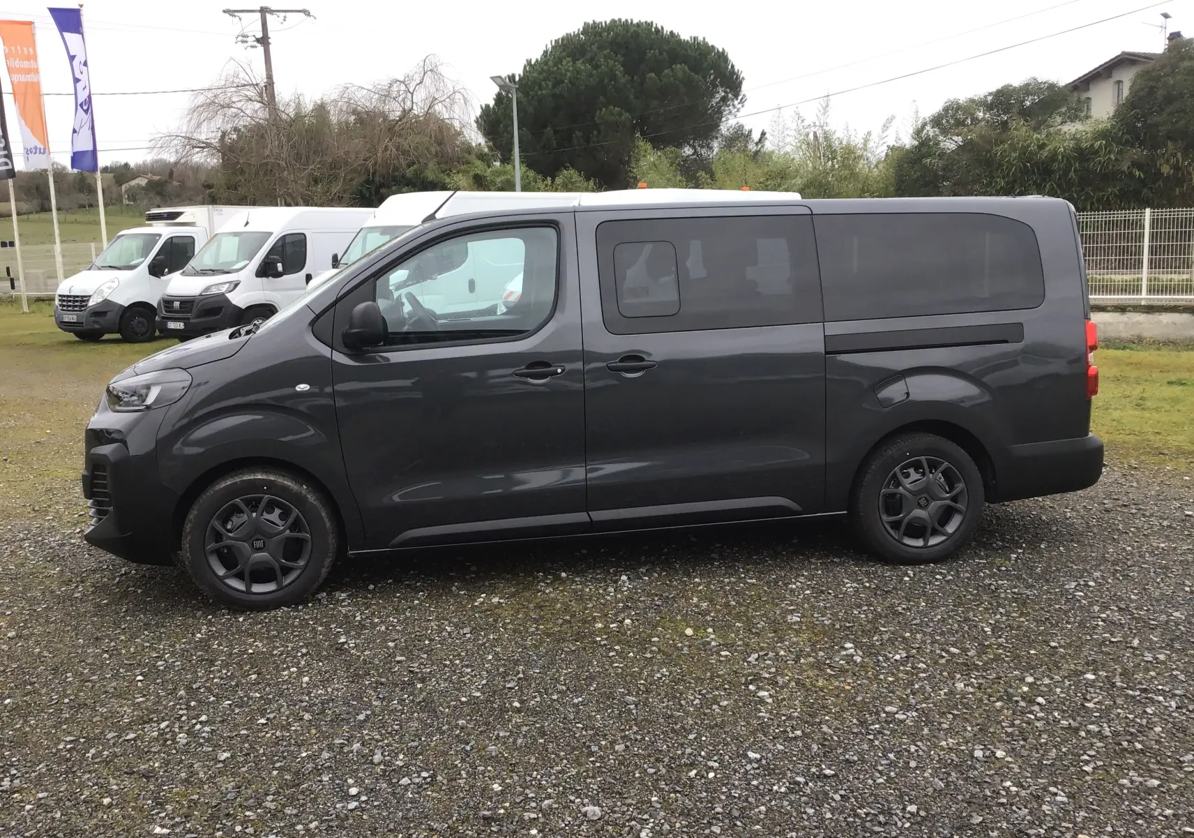 Vue de profil côté gauche du FIAT Scudo Combi gris titane avec vitres latérales coulissantes sur un sol gravillonné.