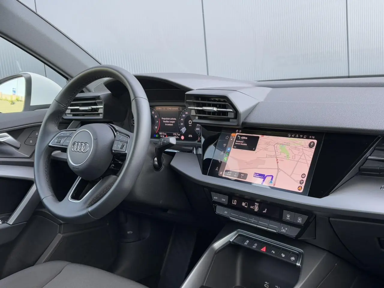 Vue intérieure côté conducteur de l'Audi A3 Sportback 2024, volant cuir et écran tactile de navigation allumé.