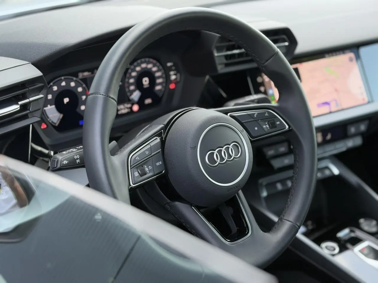 Vue rapprochée du volant cuir noir multifonctions de l'Audi A3 Sportback 2024 avec tableau de bord numérique et écran tactile.